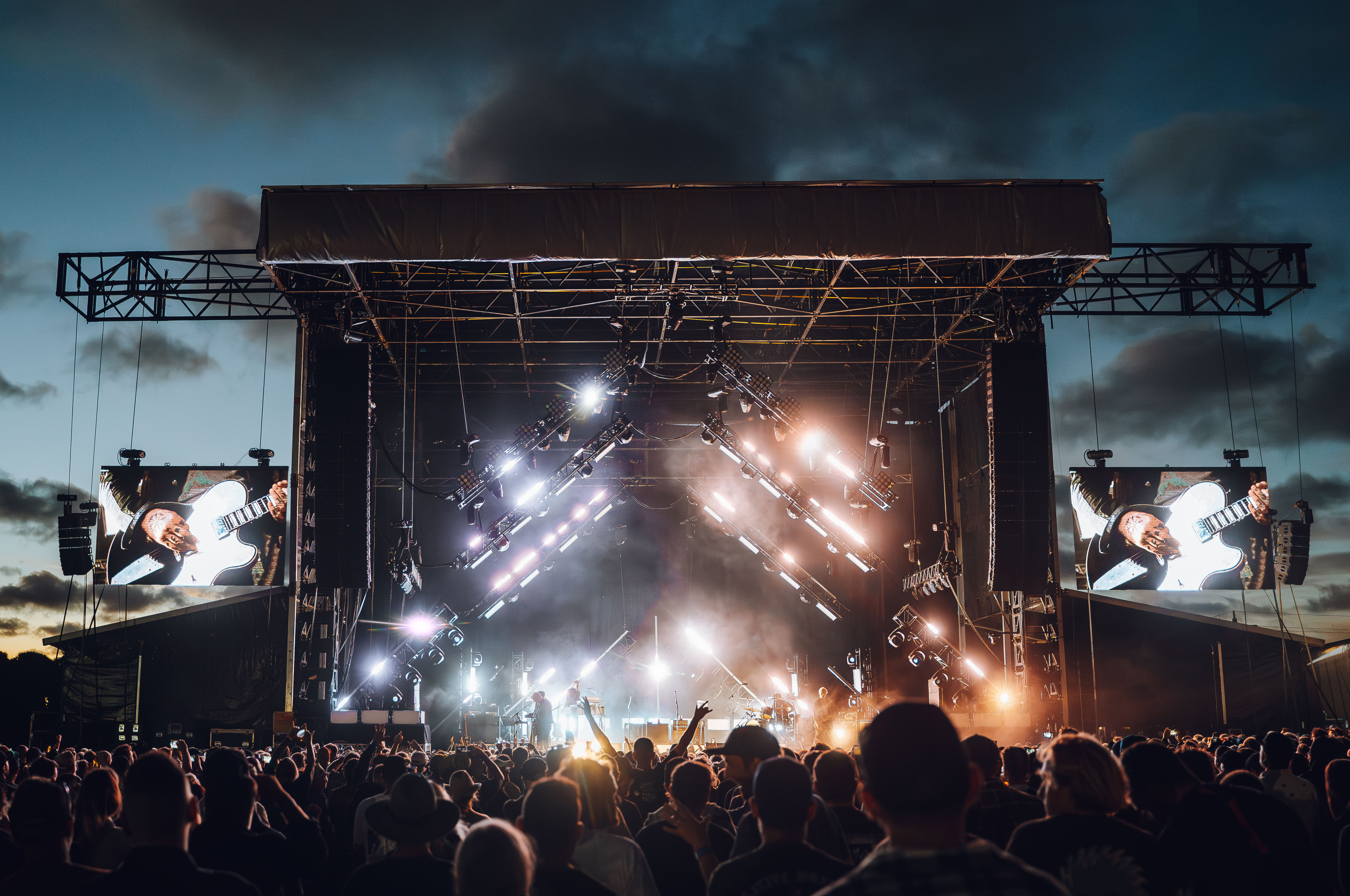 Crowd facing a floodlit outdoor concert stage at dusk.
