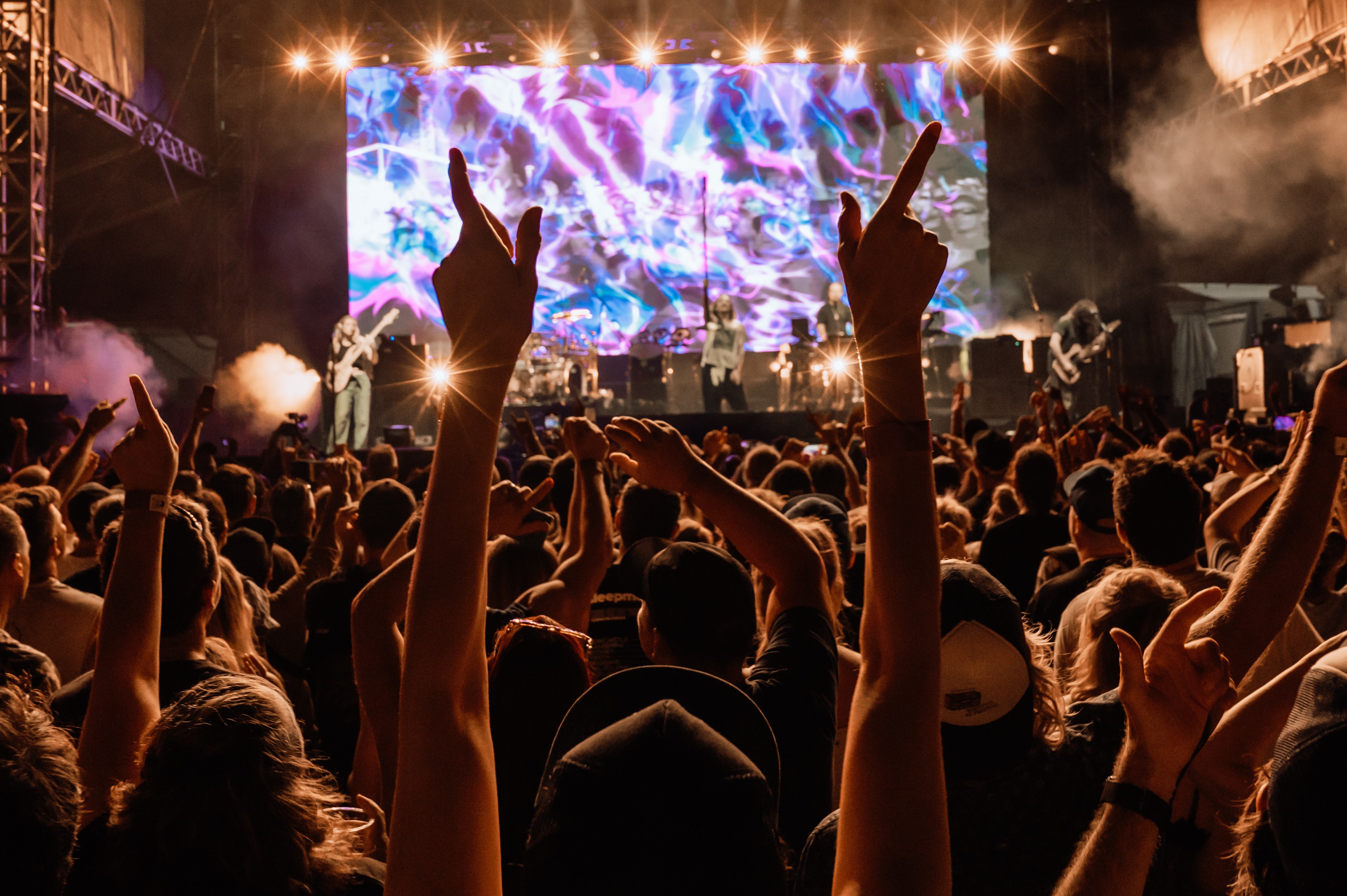 Concert crowd with raised hands in front of a lit stage screen.